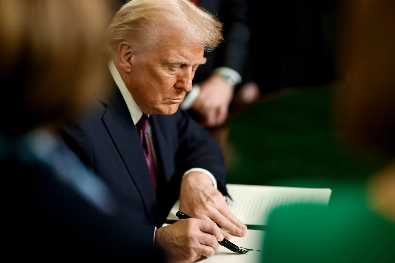 Donald Trump, assis à un bureau, signe un document avec un stylo noir.
