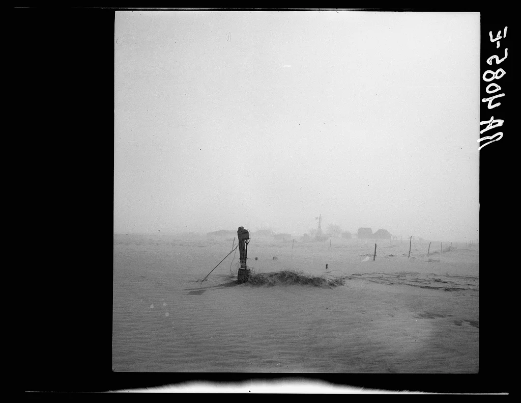Photographie en noir et blanc : le sol et les bâtiments d’une ferme disparaissent dans la poussière.