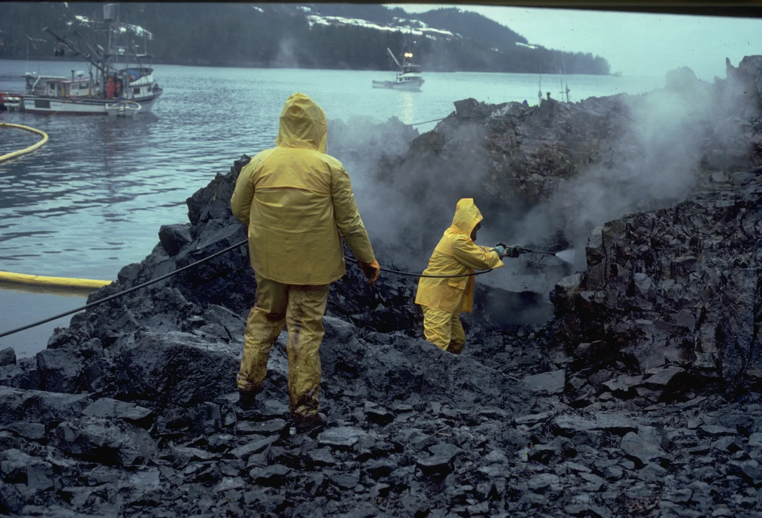 Deux personnes en imperméables jaunes nettoient des rochers noirs de pétrole au bord de l’eau.